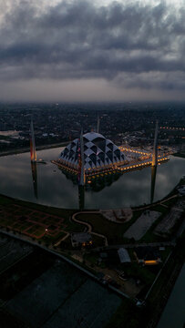 Aerial view of Al Jabbar Grand Mosque illuminated at dusk, surrounded by a reflective pond and urban landscape in Bandung, West Java, Indonesia.
