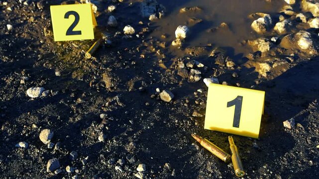 Yellow forensic evidence markers numbering one and two next to several bullet shell casings on a muddy, rocky road next to a puddle, representing a police investigation into a serious crime