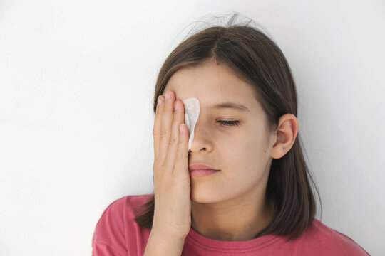 Preteen girl with brown hair in pink shirt covering eye with adhesive patch by hand, closed eyes, white wall, copy space, concept of child eye injury, pediatric ophthalmology, amblyopia treatment