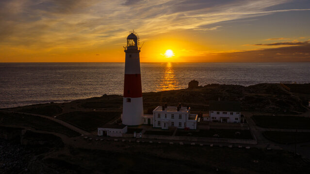 Aerial view of Portland Bill Lighthouse at sunset with the sun setting over the English Channel, rocky coastline, and calm sea in Portland, England, United Kingdom.