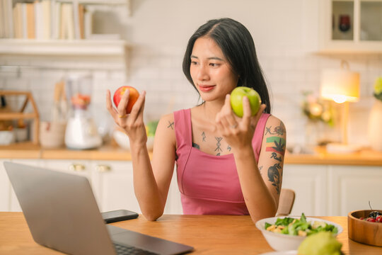 Smiling young Asian woman holding apple while working on laptop at kitchen table with salad and fresh fruit Smiling Asian woman holding apple working on laptop in home kitchen