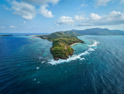 Aerial view of the lush green island surrounded by turquoise coral reefs and deep blue ocean waters under a bright sky in Karimunjawa, Central Java, Indonesia.