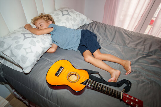 A tired toddler sleeps on a bed next to an acoustic guitar