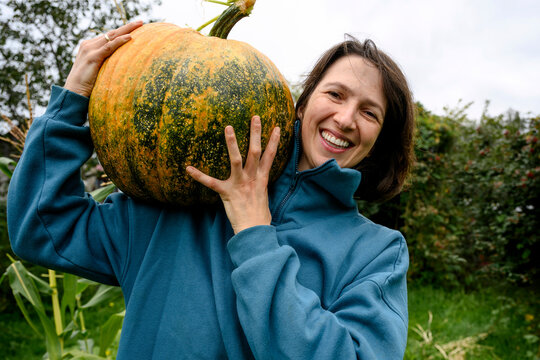 Smiling mature woman holds a large harvested pumpkin outdoors