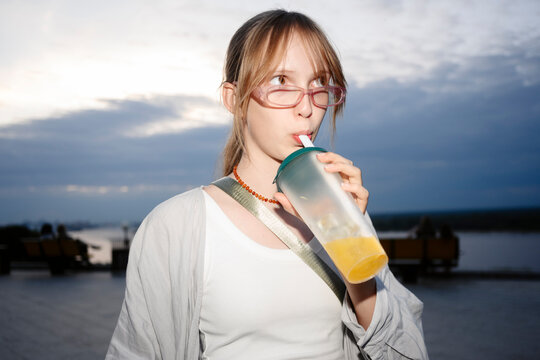A girl drinks a beverage from a plastic cup outdoors by the river