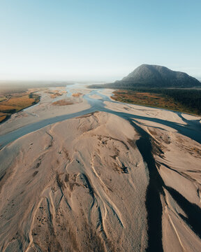 Aerial view of the braided Haast River flowing through a vast landscape with sandy riverbeds and a prominent mountain in the background in Haast, West Coast Region, New Zealand.
