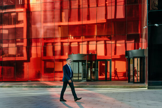 Businessman walks outdoors past a modern red glass office building