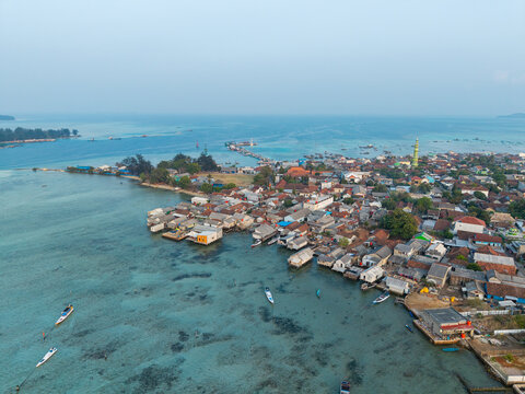 Aerial view of Karimunjawa Village with stilt houses over the turquoise sea, small boats, and a mosque minaret under a clear sky in Karimunjawa, Central Java, Indonesia.
