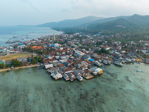 Aerial view of Karimunjawa Village with stilt houses over the clear turquoise water, traditional rooftops, and lush green hills in the background, Karimunjawa, Central Java, Indonesia.