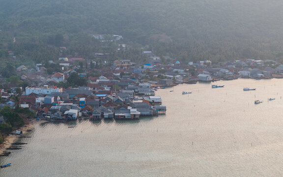 Aerial view of Karimunjawa Village with stilt houses along the coast, small fishing boats on calm water, and green hills in the background in Karimunjawa, Central Java, Indonesia.