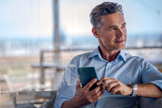 Thoughtful businessman holding smartphone by window in office
