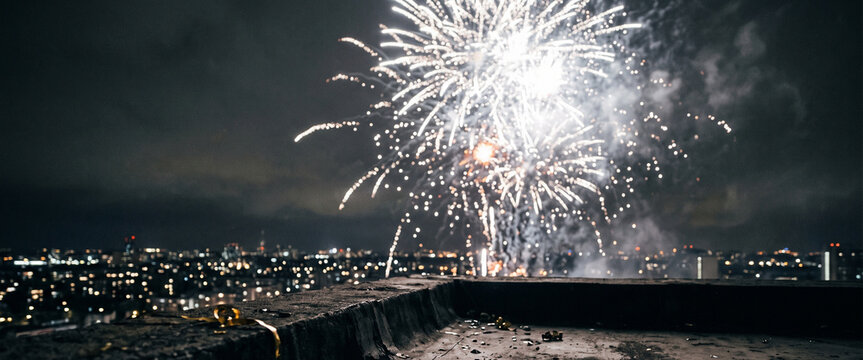 Fireworks explode in a night sky over a city skyline, illuminating the dark atmosphere and creating a festive ambiance. Captured from a rooftop perspective.