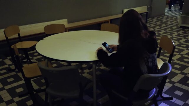 Young female student with long hair sitting alone at a round table in an empty, dimly lit school hallway. She is scrolling through social media on her smartphone, feeling isolated and lonely