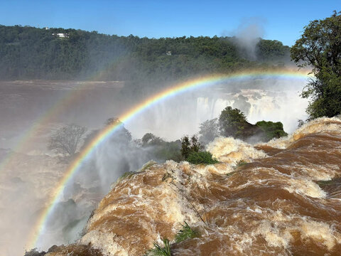 Aerial view of Iguazu Falls with a vibrant double rainbow arching over powerful rushing water, surrounded by lush rainforest in Misiones Province, Argentina.