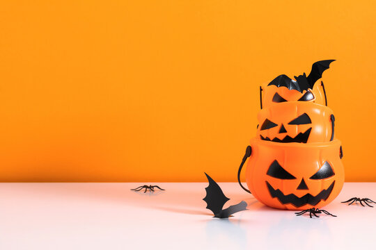 Halloween stacked jack-o-lantern pumpkin buckets with flying paper bats and plastic spiders on white table. Spooky October holiday celebration background with copy space on bright orange backdrop.