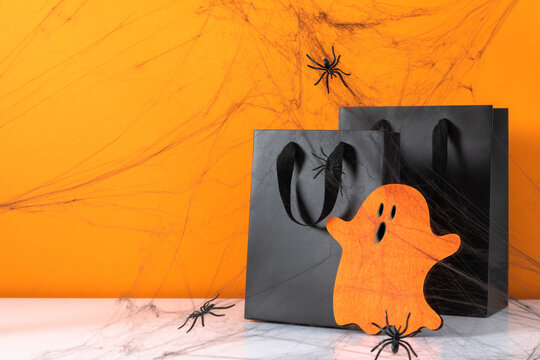 Halloween black shopping bags with cute orange ghost cutout, spiders and spooky cobweb on white table. Holiday sale and retail commercial concept background with copy space on bright orange backdrop.