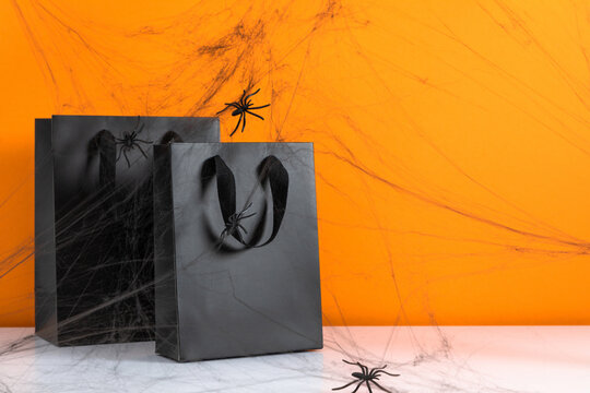 Halloween black shopping bags covered with spooky spider web and plastic spiders on white table. Creative holiday sale and commercial concept background with copy space on bright orange backdrop.