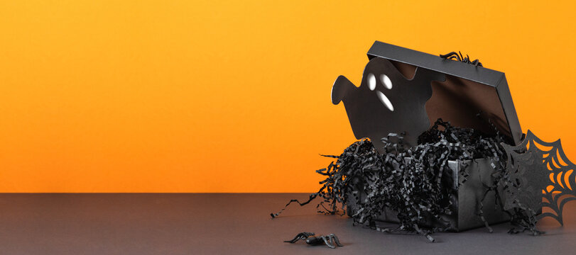 Halloween black gift box with scary ghost cutout, spiders and shredded paper. Spooky gothic present container for October holiday party celebration with copy space on bright orange background.