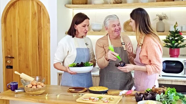 Three people in aprons cook together in a cozy home kitchen, decorating a dish with a piping bag and preparing salad and pastry beside fresh ingredients on a wooden countertop.