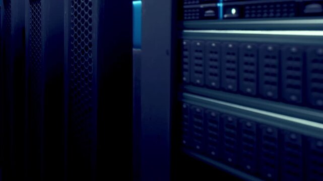 Rows of server racks stand in a dark, shadowy data center. Soft blue lights illuminate the panels, casting a futuristic atmosphere filled with quiet hums and flickering lights.