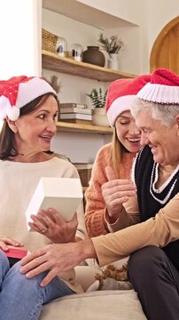 Smiling family in Santa hats exchange a gift on a cozy couch, sharing holiday cheer in a warmly decorated living room