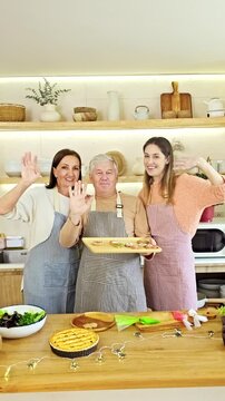 Three people in aprons cook and wave in a bright home kitchen, presenting freshly baked tarts beside a salad on a wooden countertop.