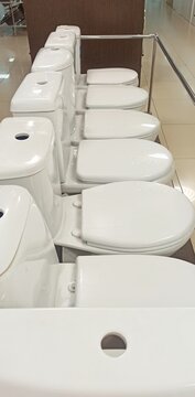 Row of toilet lids in a plumbing store. Multiple white toilet seats with open cistern holes displayed in a showroom aisle with metal railing and tiled floor, indoor retail, no people.