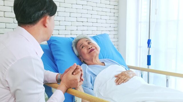 An elderly woman rests in a hospital bed, smiling as a man holds her hands, offering comfort beside an IV stand in a bright patient room.