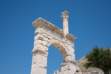 Pale stone arch rises beside a fluted column at Sagalassos. Ornate moldings and a carved capital frame broken masonry against blue sky © Ensar