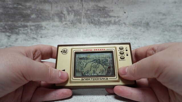 Hands holding the Soviet portable electronic liquid crystal game &ldquo;Secret Oceans". A close-up of an antique pocket electronic toy with a monochrome screen and control buttons on a gray surface.