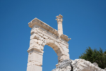 Ancient stone arch rises above foreground rocks at Sagalassos. Broken blocks, foreground rock, green leaves, and blue sky frame the low-angle view © Ensar