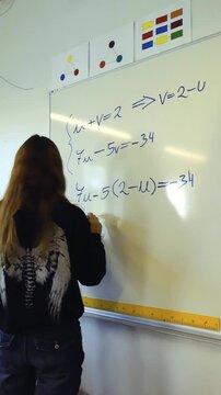 Young long haired student in a classroom writing and solving a system of linear equations with two variables on a whiteboard, demonstrating educational problem solving skills in a school setting