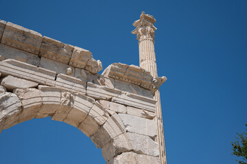An ancient stone arch and ornate column rise against blue sky at Sagalassos. A carved relief marks the center of the arch. © Ensar