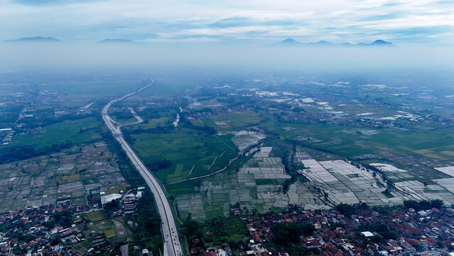 Aerial view of the Soroja Toll Road winding through lush green rice paddies and residential neighborhoods in Soreang, Bandung, West Java, Indonesia.
