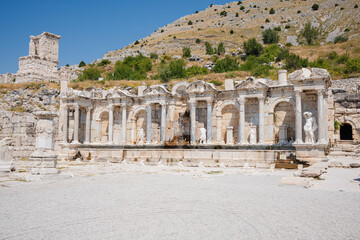 Ornate pale stone fountain facade stands amid the ruins of Sagalassos. Columns, arches, sculptures, and a stained channel face a gravel forecourt © Ensar