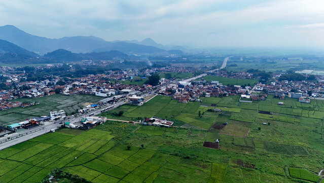 Aerial view of Soreang town surrounded by lush green rice fields, residential rooftops, and distant mountains under a soft sky in Bandung, West Java, Indonesia.