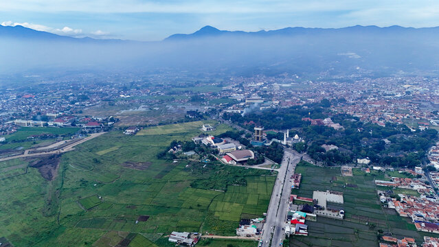 Aerial view of Soreang with its prominent mosque tower, surrounded by lush green rice fields, urban residential areas, and distant mountains in Bandung, West Java, Indonesia.