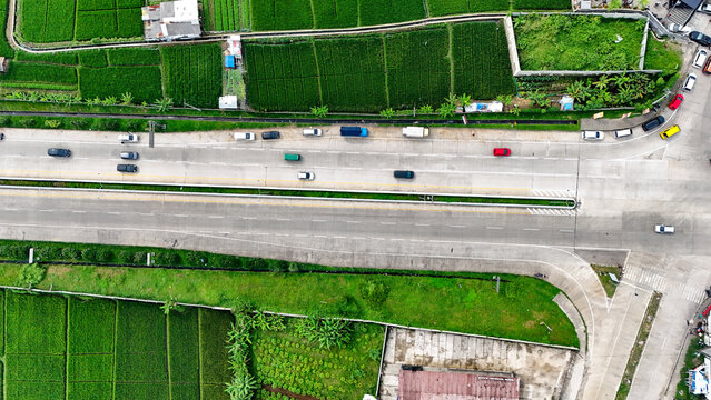 Aerial view of a highway in Soreang with cars driving along the road, flanked by lush green rice fields and agricultural land in Bandung, West Java, Indonesia.