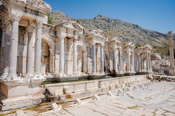 Carved pediments and Corinthian capitals crown columns above long stone fountain pool at Sagalassos in Turkey. Columns frame the ancient fountain and paved square beneath hills at ancient city © Ensar
