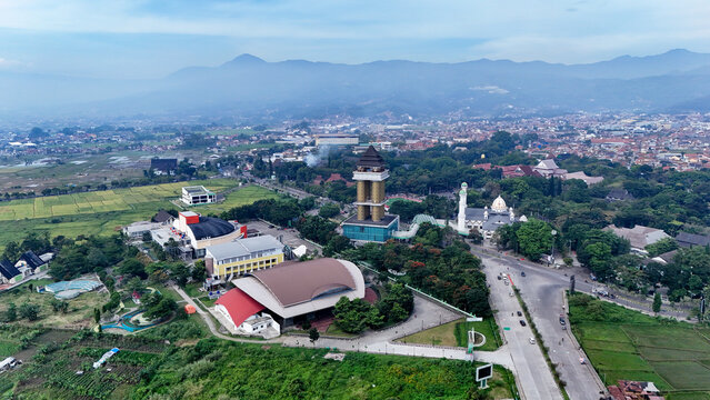 Aerial view of Soreang featuring a prominent tower, a mosque, surrounding urban buildings, and lush green rice fields under a clear sky in Bandung, West Java, Indonesia.