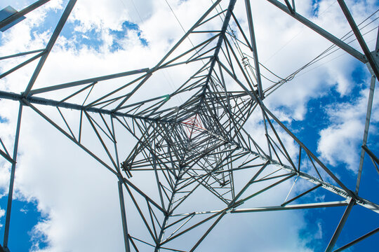 A massive high-voltage electricity pylon is shown, captured from a low angle looking upwards. Behind the metal framework, a brilliant, deep blue sky adorned with white clouds is visible. The image fea