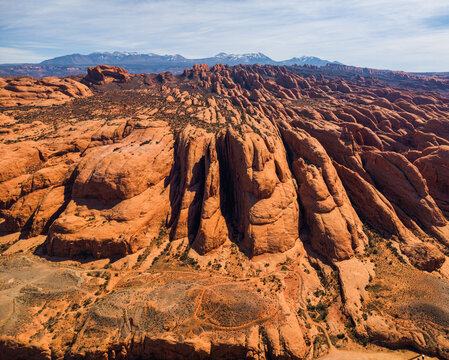 Aerial view of the rugged red rock formations and canyons along Potash Road with desert vegetation and distant snow-capped La Sal mountains in Moab, Utah, United States.