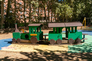 Green wooden play train with locomotive and passenger carriage stands on colorful safety surfacing beneath mature trees in Spain. Empty playground equipment represents childhood recreation, imaginativ © Volha