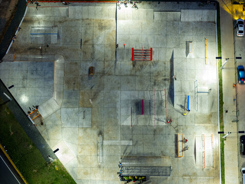 Aerial view of a concrete skatepark at night, illuminated by floodlights, featuring ramps, rails, and obstacles with people skating, in Santo Domingo, Distrito Nacional, Dominican Republic.