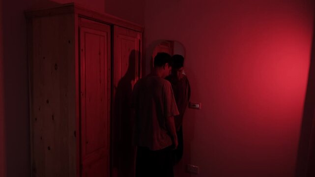 Man staring at his reflection before lowering his head and resting against the mirror, expressing self rejection, hopelessness, shame and emotional despair in red low key light