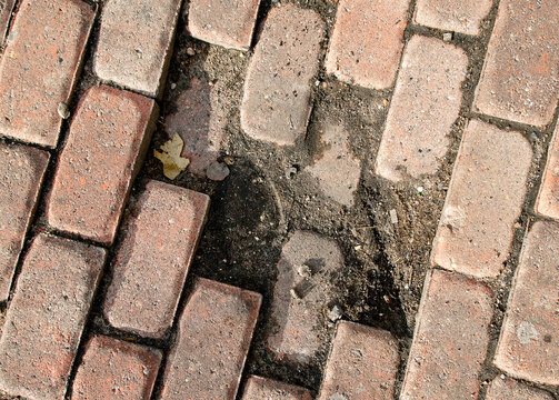Broken pavement blocks on an urban walkway, missing red pavers creating trip hazard on sidewalk surface. Damaged red brick paving on city sidewalk, missing stones requiring maintenance repair