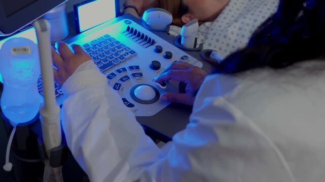 Close up view of a medical specialist's hands using an ultrasound machine's keyboard and trackball during a patient examination in a dark room with blue lighting.