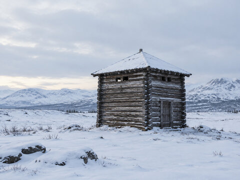 Square log blockhouse isolated in a snow-covered wilderness, soft winter light, desolate landscape.