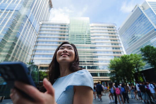 Smiling woman holds smartphone and looks up at city skyscrapers