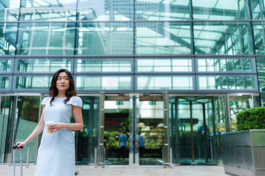 A businesswoman holds coffee and luggage outside a corporate building